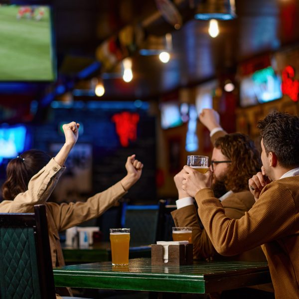 Close-up of friends group toasting with beer while sitting in beer pub together. Selective focus on man holding mug with cool drink